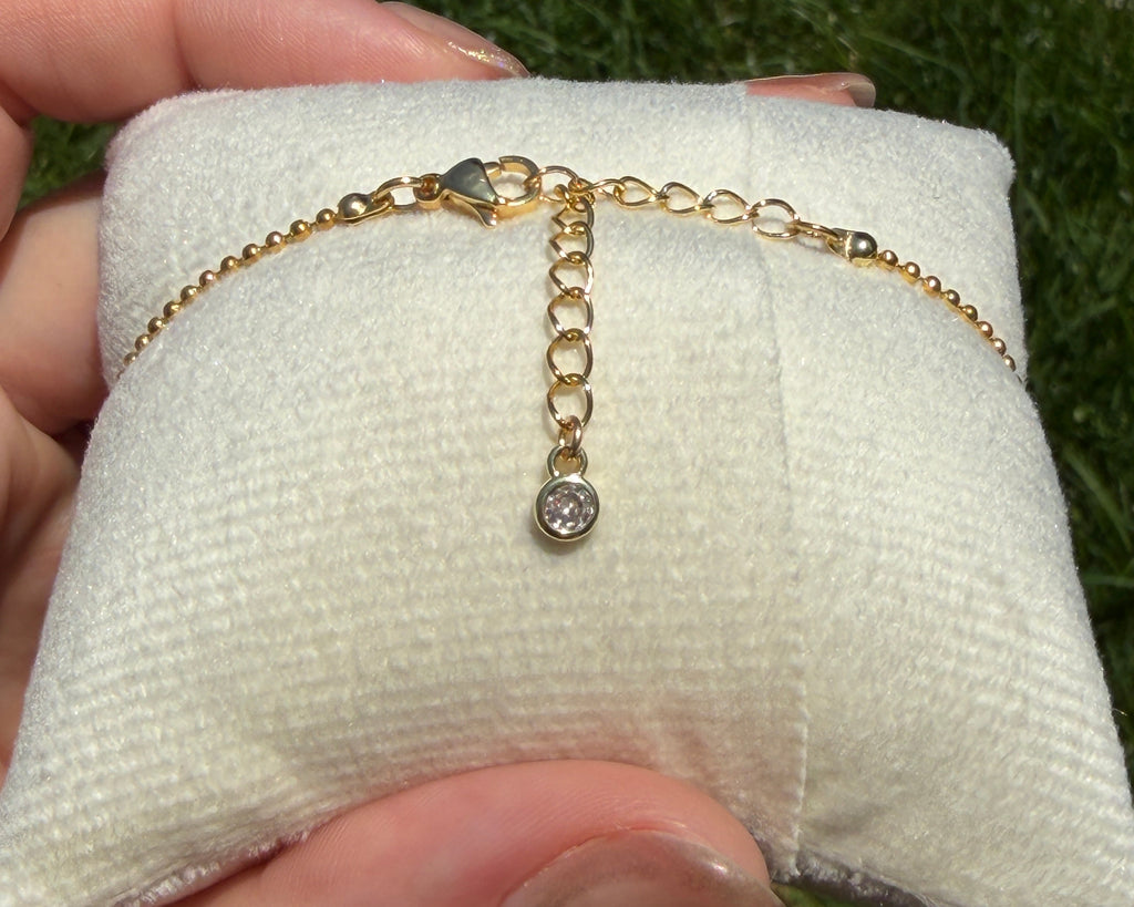 Gold bracelet with a small gemstone on a white cushion, held by a hand against a grassy background.